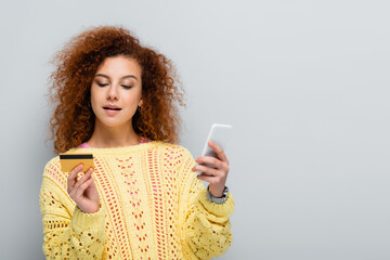 Wall Mural - curly woman in knitted sweater holding smartphone and credit card isolated on grey