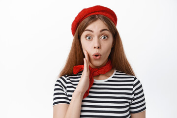 Wall Mural - Close up of excited young girl in red beret and scarf, telling big secret or news, holding hand near mouth, talking to you, making announcement, standing over white background