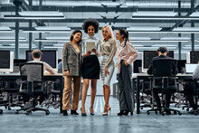 A Group Of Young Successful Business Women Working Together Standing In The Middle Of The Office And Looking At A Tablet. In The Background Are Colleagues Working On Computers.