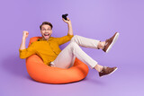 Fototapeta Panele - Full length body size photo of guy sitting in beanbag playing video games with controller gesturing like winner isolated on vivid violet color background