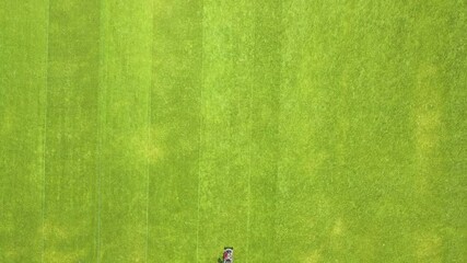 Wall Mural - Aerial view of small figure of man worker trimming green grass with mowing mashine on football stadium field in summer.