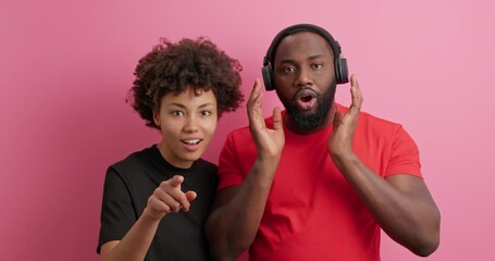 Wall Mural - Afro American woman interrupts boyfriend who listens music via headphones points forward shows something amazing say wow impressed by incredible thing isolated over pink background. Hey look there