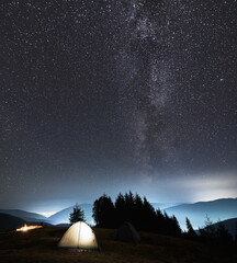 Two tourist tents set up on mountain hill under night starry sky against background of magical Milky Way, shadows of trees and contours of the mountain beskids. Concept of night camping and astronomy.