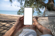 © marchsirawit - Hand holding mock up mobile with white sceen while laying on hammock swing between trees on the beach.