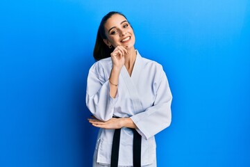 Wall Mural - Beautiful brunette young woman wearing karate fighter uniform with black belt looking confident at the camera with smile with crossed arms and hand raised on chin. thinking positive.