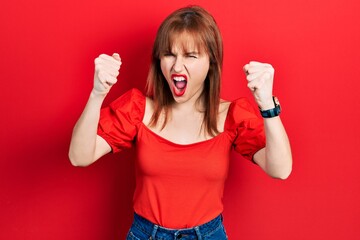 Canvas Print - Redhead young woman wearing casual red t shirt angry and mad raising fists frustrated and furious while shouting with anger. rage and aggressive concept.