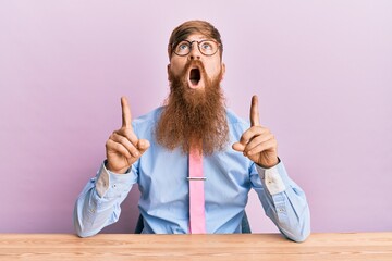 Wall Mural - Young irish redhead man wearing business shirt and tie sitting on the table amazed and surprised looking up and pointing with fingers and raised arms.