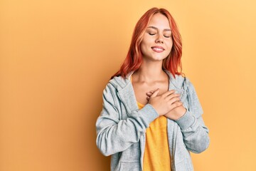 Canvas Print - Young beautiful redhead woman wearing casual sporty sweatshirt smiling with hands on chest with closed eyes and grateful gesture on face. health concept.