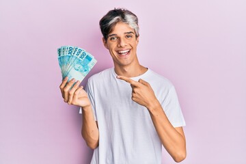 Poster - Young hispanic man holding 100 brazilian real banknotes smiling happy pointing with hand and finger