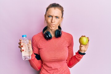 Canvas Print - Beautiful caucasian sports woman holding water bottle and green apple skeptic and nervous, frowning upset because of problem. negative person.