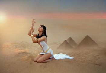 Wall Mural - Egypt Style Woman. Sexy beautiful girl goddess Queen Cleopatra sits on sand desert pyramids. Art ancient pharaoh costume white dress gold accessories. Egyptian makeup. hands raised to sky lady praying