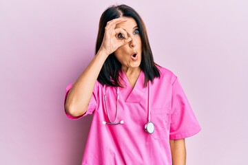 Canvas Print - Middle age brunette woman wearing doctor uniform and stethoscope doing ok gesture shocked with surprised face, eye looking through fingers. unbelieving expression.