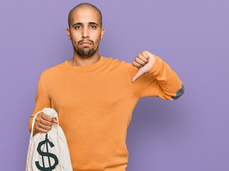 Poster - Hispanic adult man holding dollars bag with angry face, negative sign showing dislike with thumbs down, rejection concept