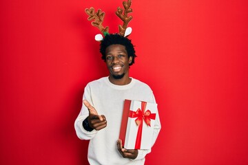 Poster - Young african american man wearing deer christmas hat holding gift pointing fingers to camera with happy and funny face. good energy and vibes.