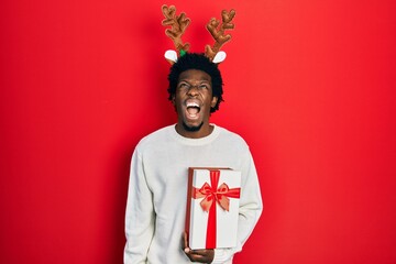 Poster - Young african american man wearing deer christmas hat holding gift angry and mad screaming frustrated and furious, shouting with anger. rage and aggressive concept.