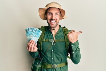 Canvas Print - Handsome man with beard wearing explorer hat holding 100 brazilian reals pointing thumb up to the side smiling happy with open mouth