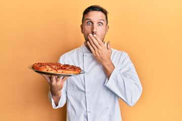 Canvas Print - Handsome man with beard professional cook holding italian pizza covering mouth with hand, shocked and afraid for mistake. surprised expression