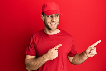 Canvas Print - Young caucasian man wearing delivery uniform and cap smiling and looking at the camera pointing with two hands and fingers to the side.