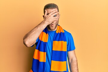 Wall Mural - Young caucasian man football hooligan cheering game peeking in shock covering face and eyes with hand, looking through fingers afraid