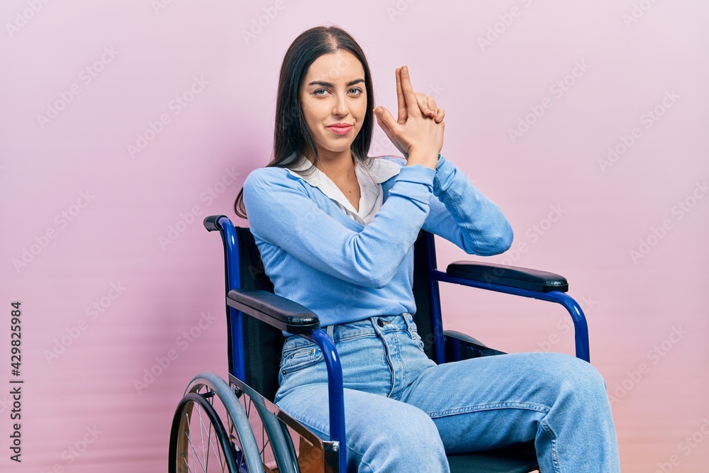 Beautiful woman with blue eyes sitting on wheelchair holding symbolic gun with hand gesture, playing killing shooting weapons, angry face