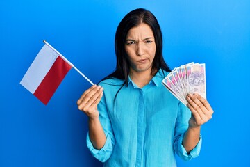 Poster - Young latin woman holding poland flag and zloty banknotes skeptic and nervous, frowning upset because of problem. negative person.