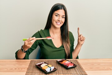 Wall Mural - Young brunette woman eating sushi sitting on the table smiling with an idea or question pointing finger with happy face, number one