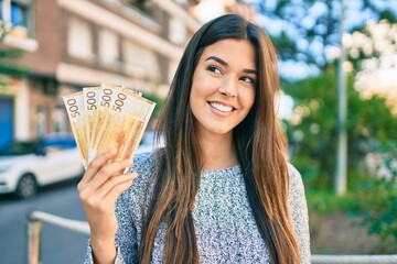 Poster - Young beautiful hispanic girl smiling happy holding norwegian krone banknotes at the city.