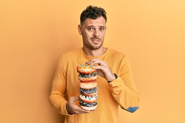 Wall Mural - Young hispanic man holding tasty colorful doughnuts clueless and confused expression. doubt concept.