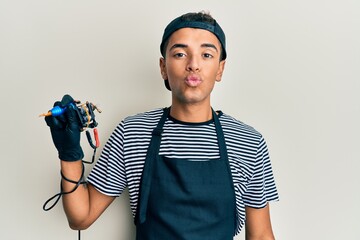 Poster - Young handsome african american man tattoo artist wearing professional uniform and gloves holding tattooer machine looking at the camera blowing a kiss on air being lovely and sexy. love expression.