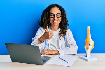 Poster - Beautiful middle age woman doctor at orthopedic clinic doing happy thumbs up gesture with hand. approving expression looking at the camera showing success.