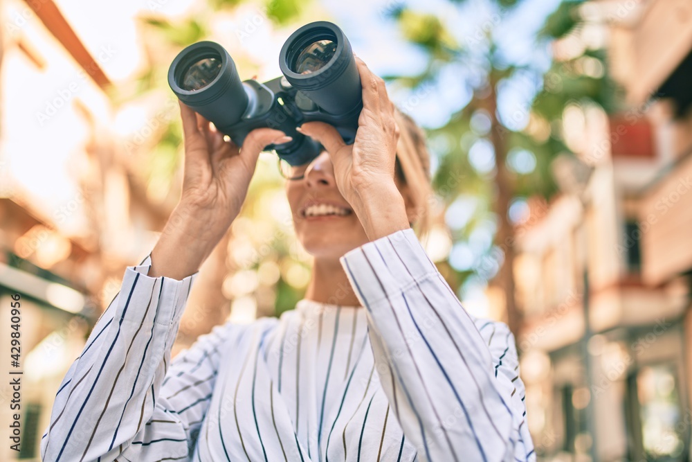 Young blonde businesswoman smiling happy using binoculars at the city.