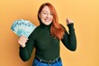 Beautiful redhead woman holding 100 brazilian real banknotes screaming proud, celebrating victory and success very excited with raised arm
