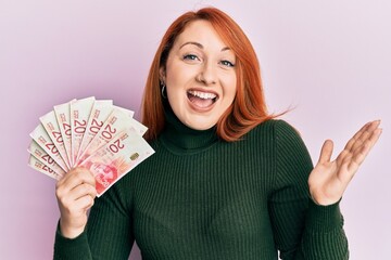 Wall Mural - Beautiful redhead woman holding 20 israel shekels banknotes celebrating achievement with happy smile and winner expression with raised hand