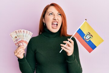 Poster - Beautiful redhead woman holding colombia flag and 10 colombian pesos banknotes angry and mad screaming frustrated and furious, shouting with anger. rage and aggressive concept.