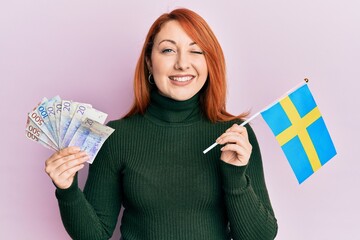 Poster - Beautiful redhead woman holding 20 swedish krona banknotes and sweden flag winking looking at the camera with sexy expression, cheerful and happy face.