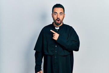 Wall Mural - Handsome hispanic man with beard wearing catholic priest robe surprised pointing with finger to the side, open mouth amazed expression.