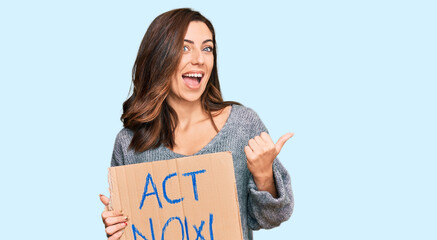 Wall Mural - Young brunette woman holding act now banner pointing thumb up to the side smiling happy with open mouth