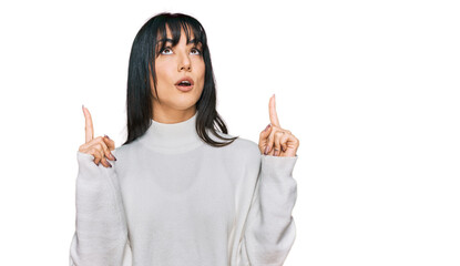 Young brunette woman with bangs wearing casual turtleneck sweater amazed and surprised looking up and pointing with fingers and raised arms.