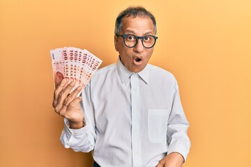 Sticker - Middle age indian man holding 100 new taiwan dollars banknotes scared and amazed with open mouth for surprise, disbelief face