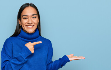 Wall Mural - Young asian woman wearing casual winter sweater amazed and smiling to the camera while presenting with hand and pointing with finger.