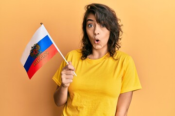 Young hispanic woman holding russia flag scared and amazed with open mouth for surprise, disbelief face