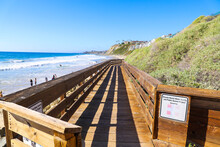 A Long Brown Wooden Bridge Along A Hillside With Lush Green Plants And Flowers With People Walking Along The Beach, Blue Ocean Water And Waves With Blue Sky At Dana Strands Beach In Dana Point, Ca
