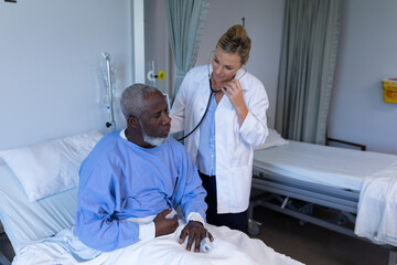 Wall Mural - Caucasian female doctor examining with stethoscope african american male patient in hospital room