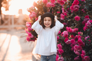 Wall Mural - Cute stylish child girl 5-6 year old wear trendy clothes and hat posing over flower rose outdoors in park. Having fun. Childhood. Happiness.