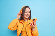 © wayhome.studio  - Horizontal shot of pretty Asian woman gets message in social networks focused at smartphone listens music via wireless headphones while having walk wears orange jacket isolated over blue wall