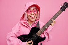 Positive Young Woman Keen On Music Plays Favorite Music Holds Electric Guitar Smiles Happily Being In Good Mood Wears Anorak With Hood On Head Trendy Sunglasses Isolated Over Pink Background
