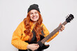 © WHstudio Leushin N - Happy redhead female guitarist plays electric guitar being in good mood being famous rock musician singer or performer wears stylish jacket and hat isolated over white background. Hobby concept