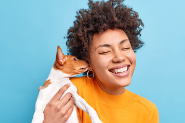 Poster - Positive Afro American woman adores cute little dog which licks her ear expresses love to owner have friendly relationships. Glad female plays with favorite pet wears orange jumper isolated on blue