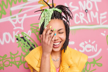 Cheerful Milennial Girl Laughs Sincerely Makes Face Palm Feels Very Happy Has Trendy Hairstyle Dressed In Casual Clothes Poses Against Colorful Graffiti Wall. Happy Female Tagger At Urban Place