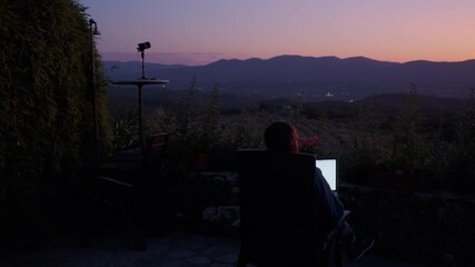 Camera panning around a man sitting in his chair and working on a laptop for work, beautiful orange sunset in the background behind mountains.